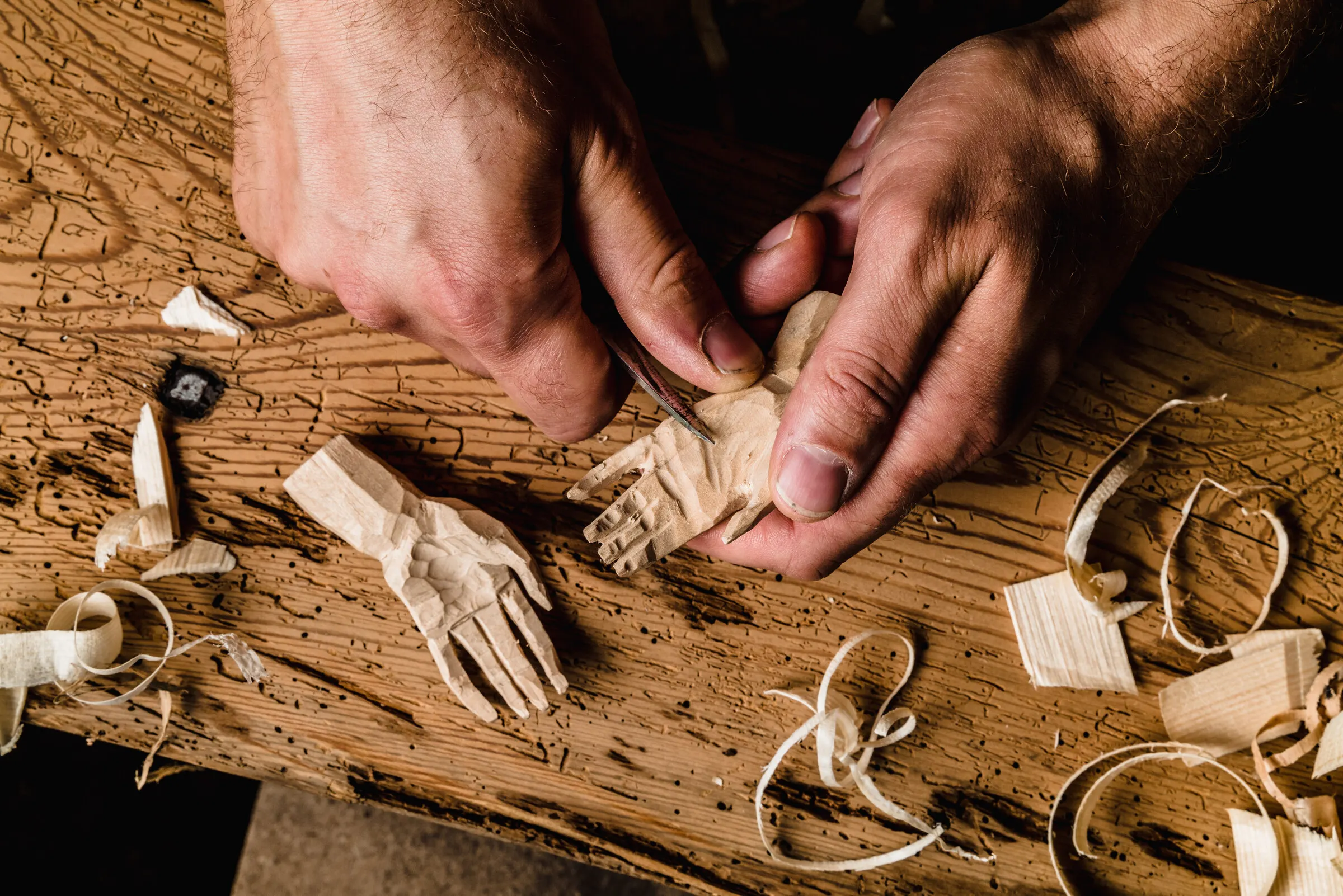 Zwei Hände schnitzen eine kleine Hand aus hellem Holz, auf einer wurmstichigen alten Bohle liegen Späne