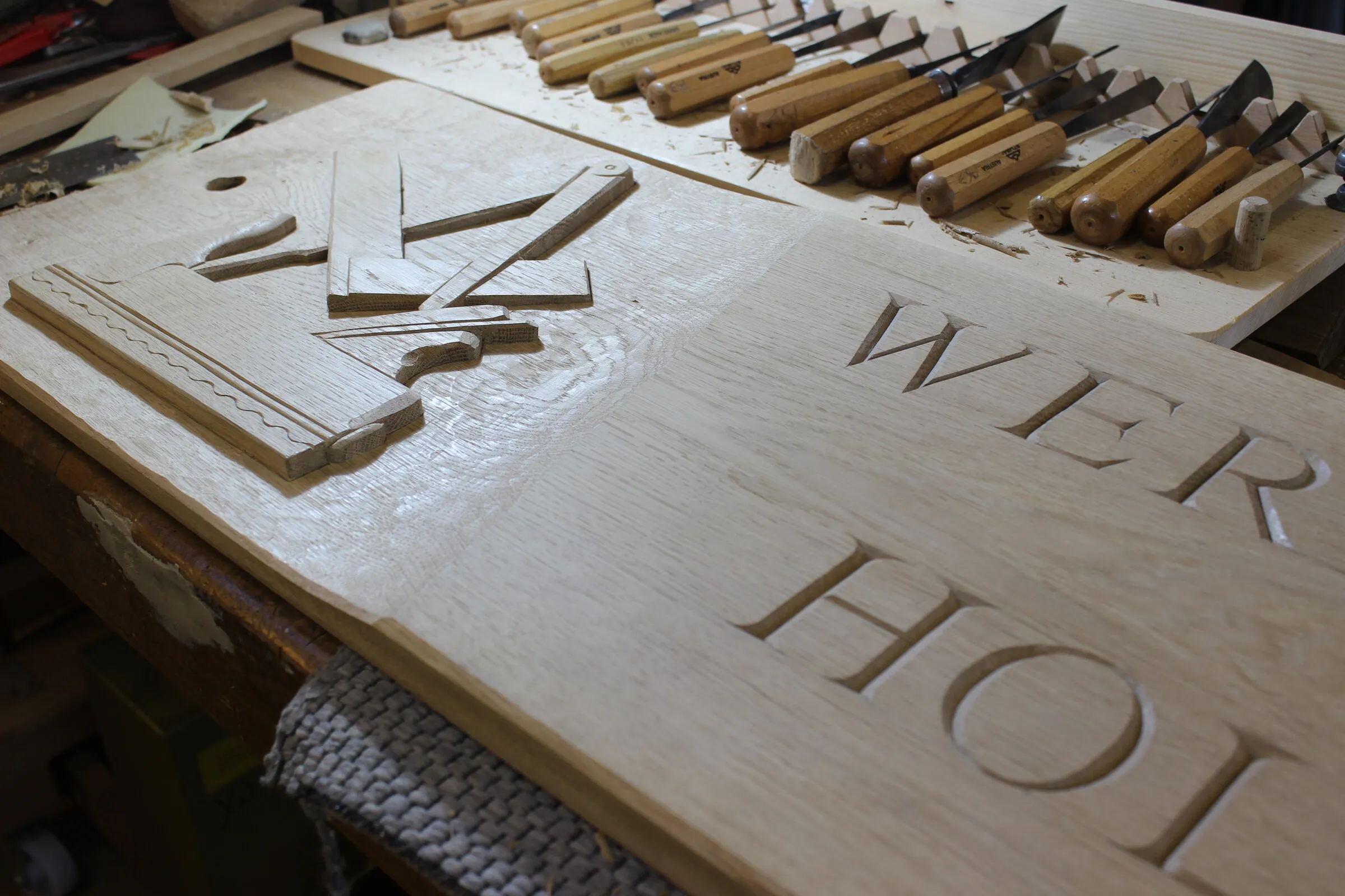 Werkstattschild in Arbeit: geschnittene Antiqua-Buchstaben in hellem Holz, daneben die Reihe der Schnitzeisen