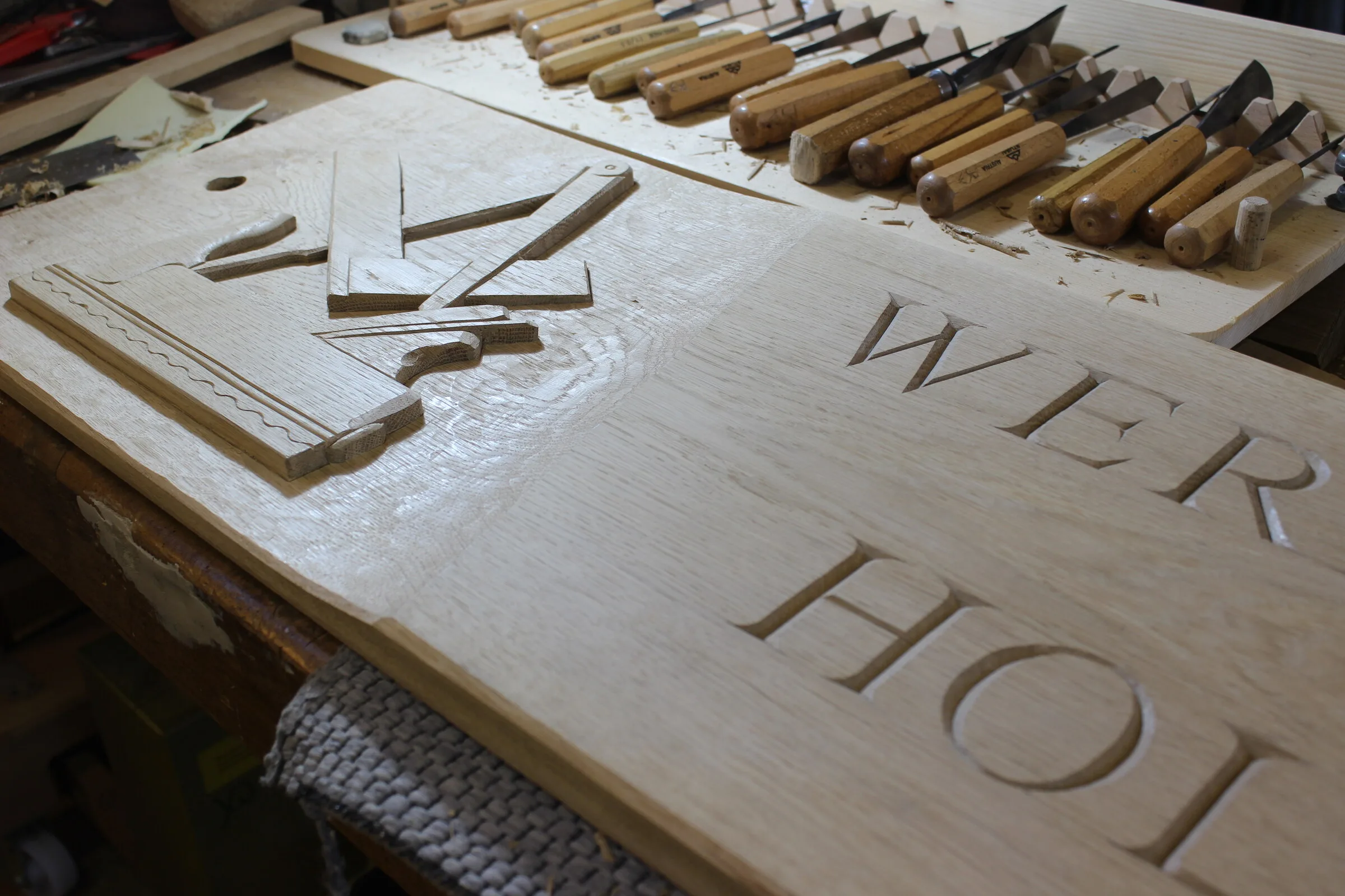 Werkstattschild in Arbeit: geschnittene Antiqua-Buchstaben in hellem Holz, daneben die Reihe der Schnitzeisen